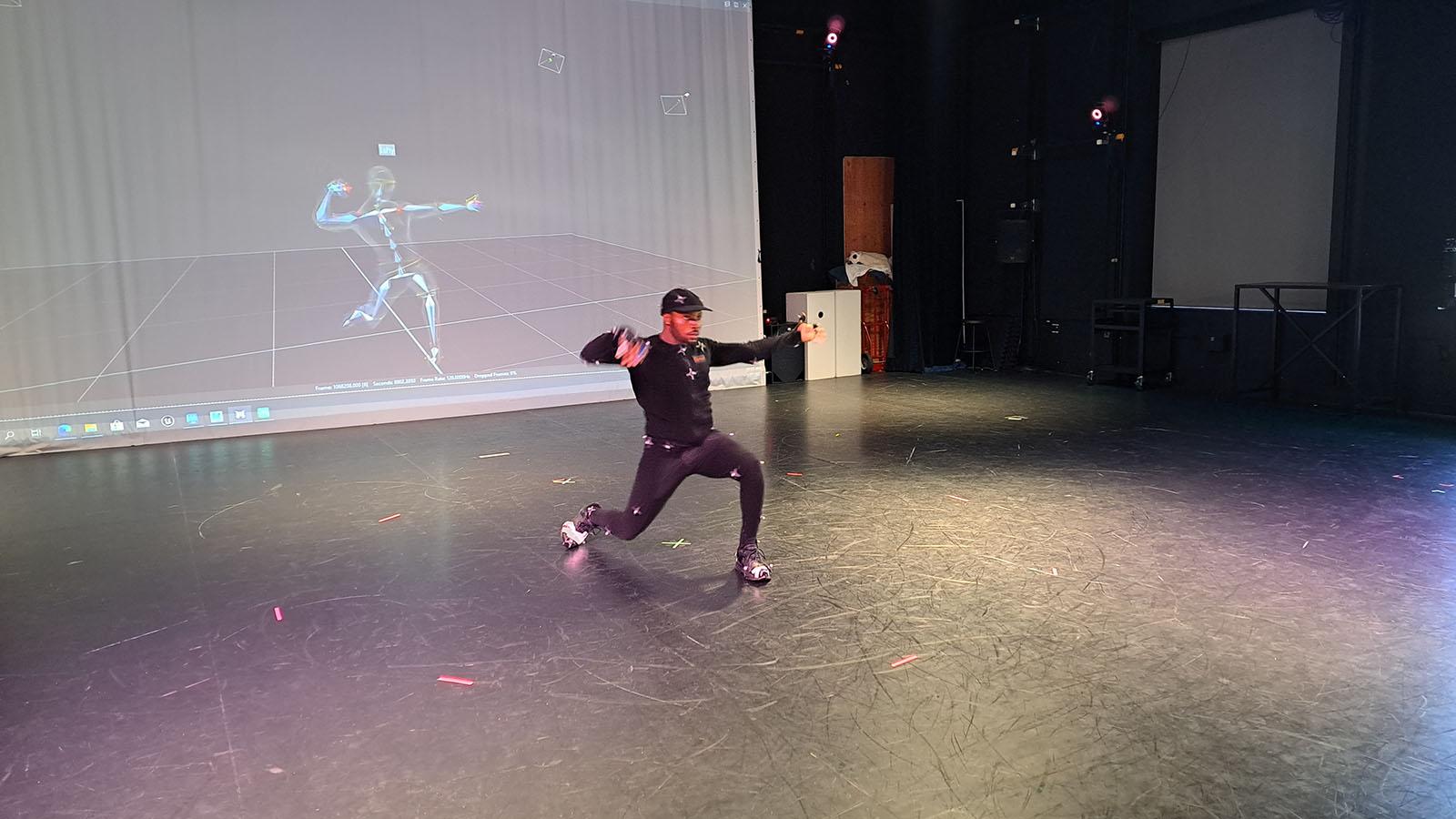 A black male dancers lunges in a mocap suit with a projection of the motion capture data on a screen behind her 