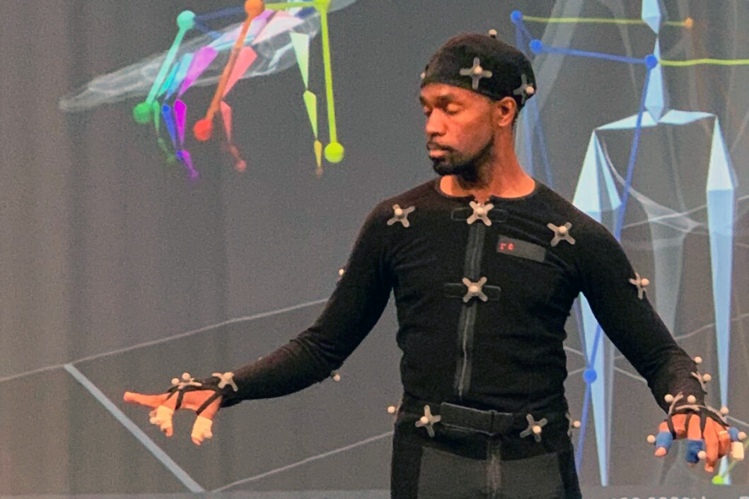 André Zachery suited up in the ACCAD Motion Lab. He looks down at the motion trackers on his fingers while his avatar is partially in view in the background.