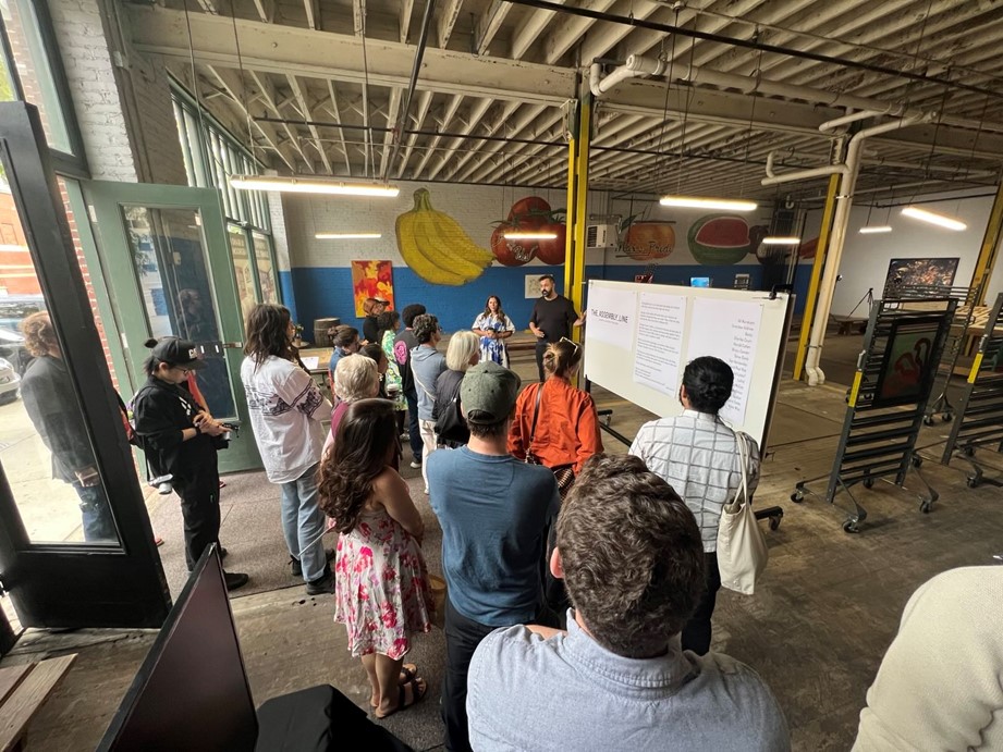 A high-angle shot from behind a group of students and faculty standing in an open, industrial gallery space with exposed ceiling beams and colorful fruit murals on the back wall. The group is facing forward, listening to two curators standing next to a large whiteboard displaying text about "The Assembly Line" exhibition.