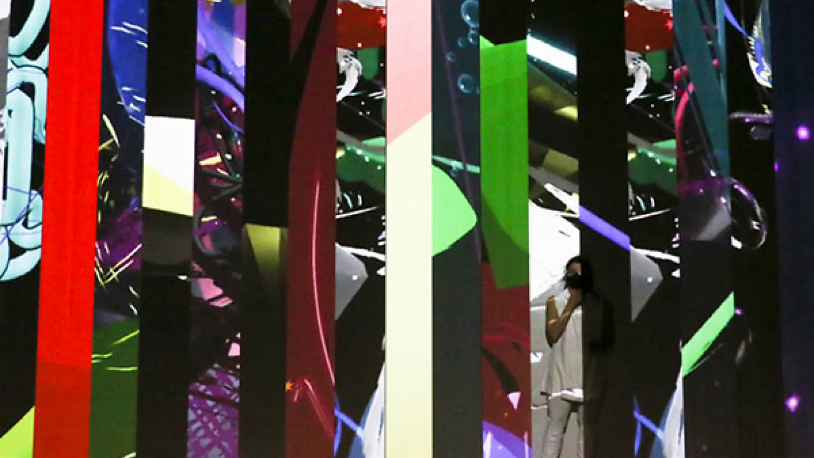 A dancer dressed white stands in front of a screen with many vertical slices of different video projections fill the space 
