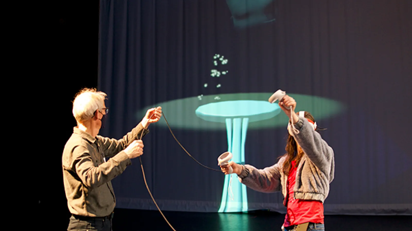 A man holds the cord of a VR headset while a person in a red shirt and gray sweatshirt experiences a VR dance experience while the animation is projected onto a screen in the background