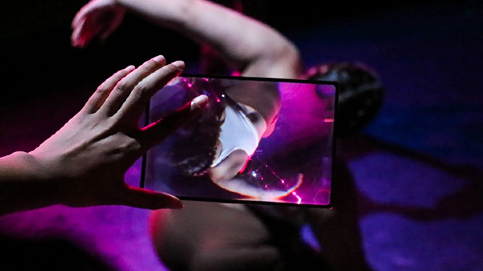 A hand holds a small plexiglass frame in front of a dancer who is spiraling their body. The frame flips and distorts the dancers image