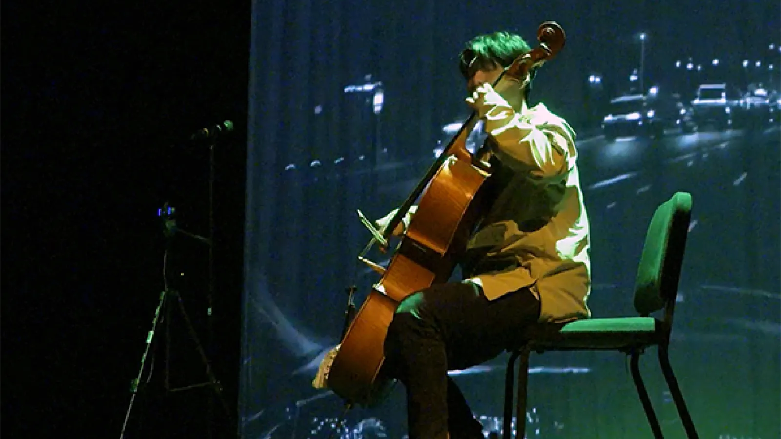 A man sitting on a chair playing a cello against a backdrop of a screen with a cityscape projected onto it.