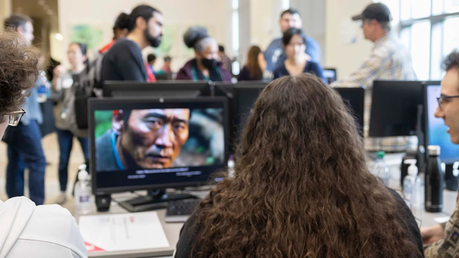 A group of three students sit at a table, watching a student-made video on a desktop computer. In the background, people socialize in the ACCAD computer lab during the Open House.