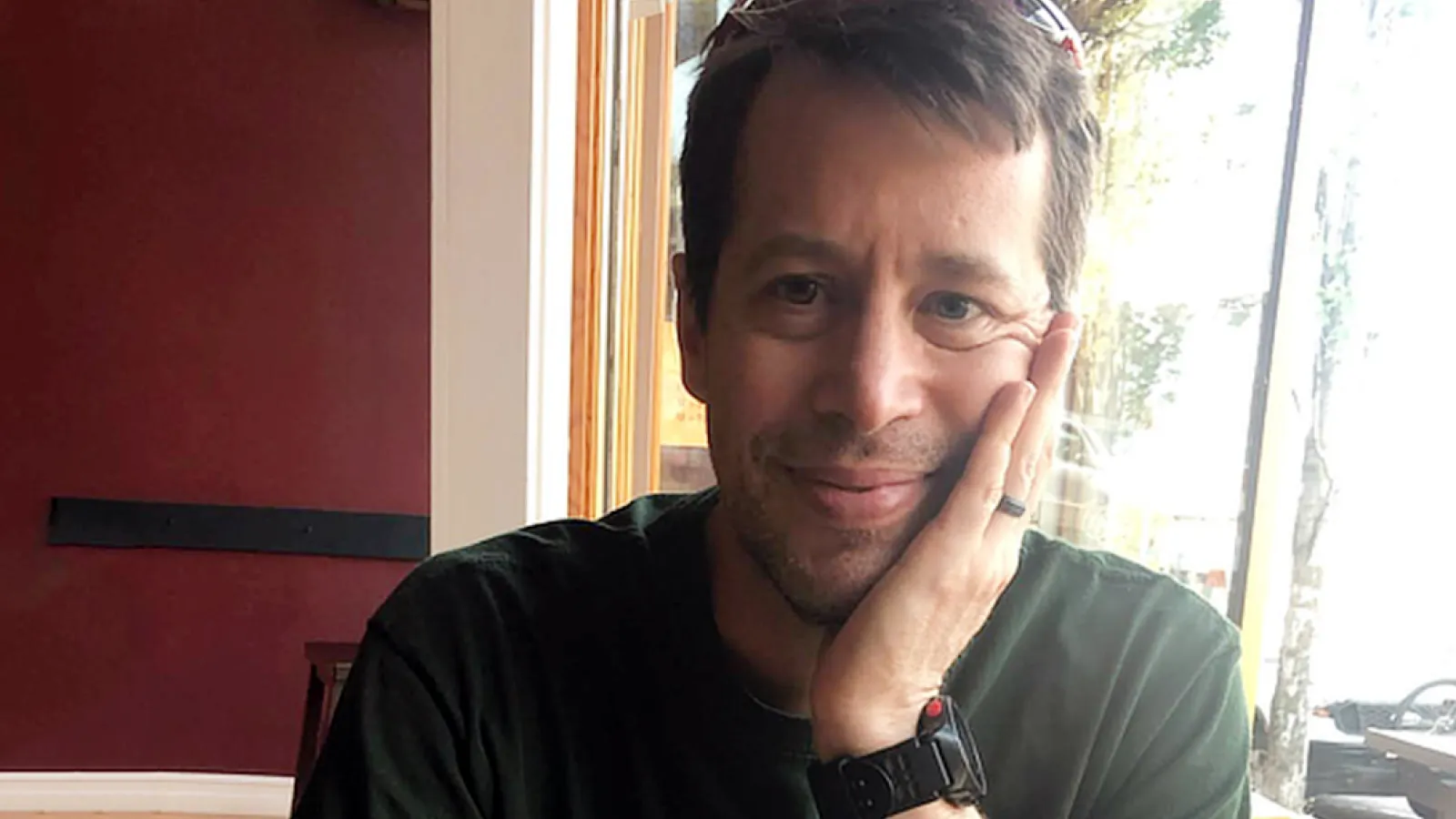 A photo of Peter Stuart, a man with short brown hair wearing a green t-shirt, and resting his head on his hand while smiling at the camera.