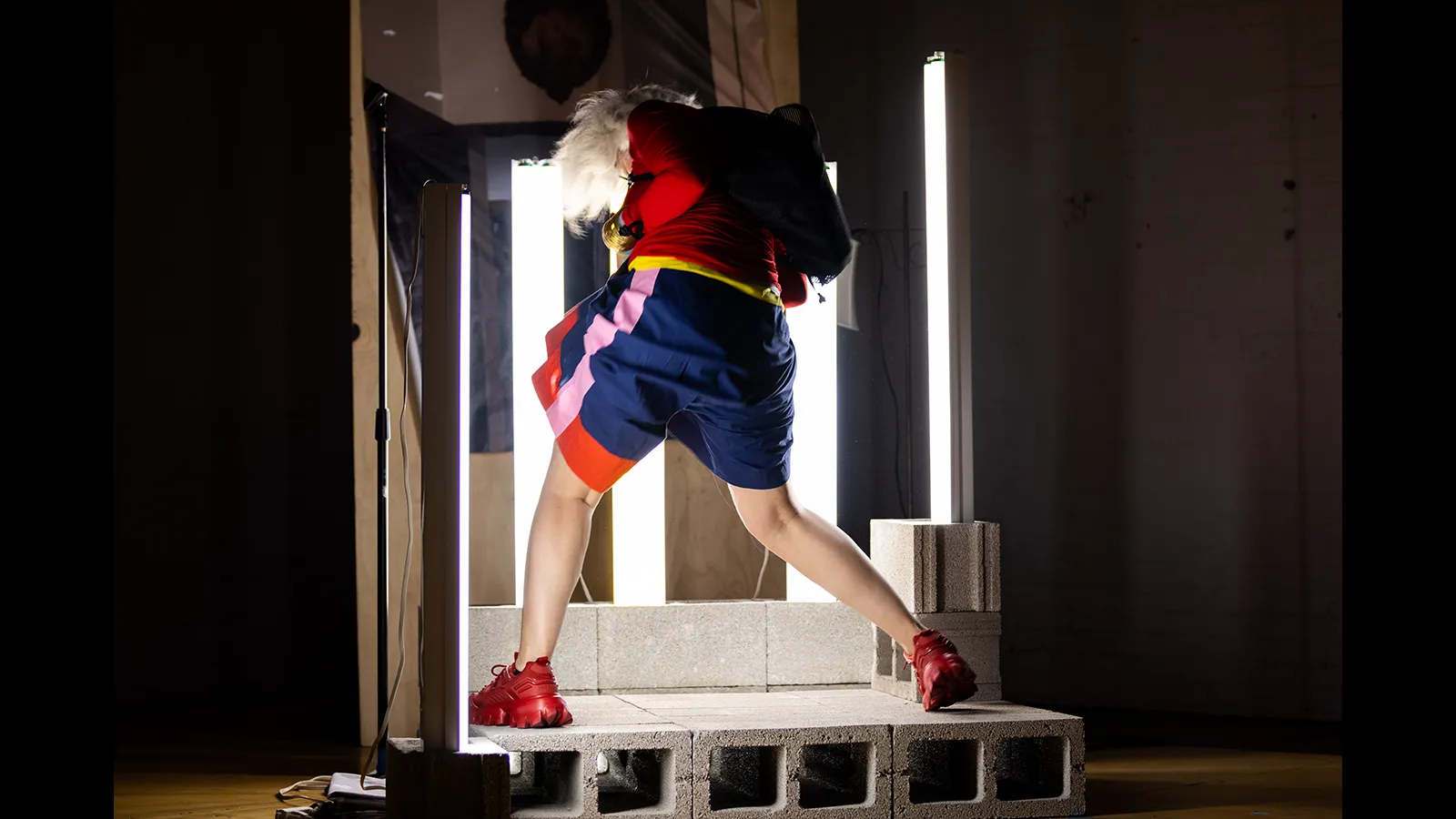 Performer moves among illuminated light tubes and cinder blocks in a dark performance space.