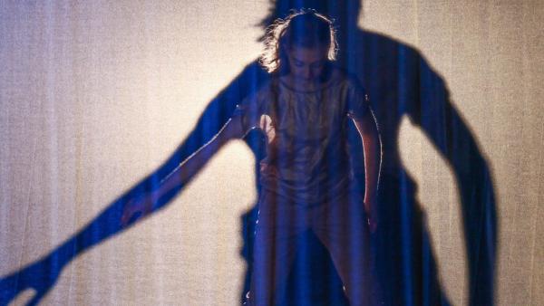 A dancer with blond hair gazes down to the floor with her right arm extended at a 45 degree angle while a larger shadow of her silhouette is projected onto a white scrim 