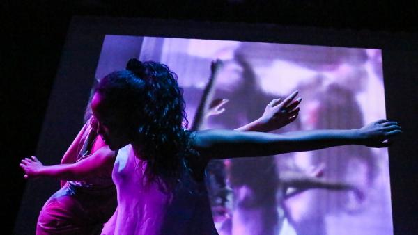 Dancers windmill their arms in front of a video loop with a soft purple wash across the screen 