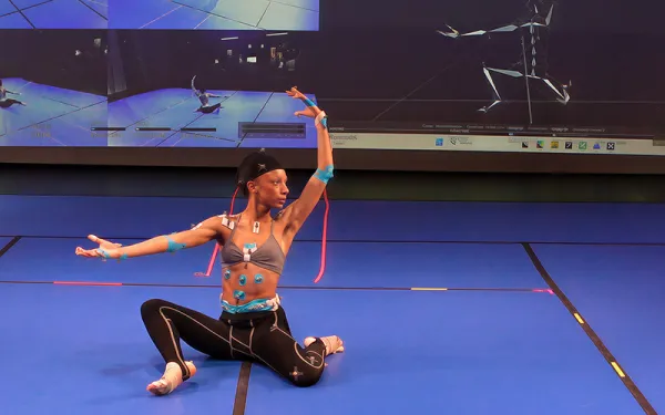 A performer wearing motion-capture sensors poses on a studio floor while large screens display a digital skeletal model and live movement-tracking data.