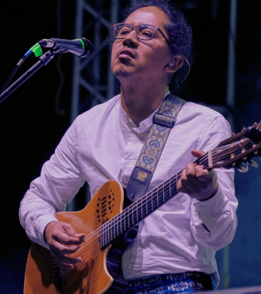 An action shot of Ati playing the guitar while standing on stage during a performance and singing into a microphone.