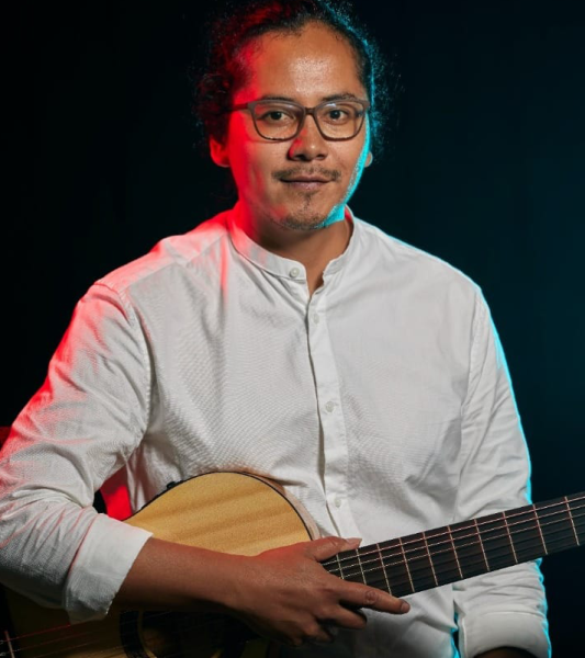 A headshot photo of Ati Cachimuel wearing a white shirt, glasses, and holding a guitar, against a black background with bright pink lighting shining in from the left and blue lighting coming in from the right.