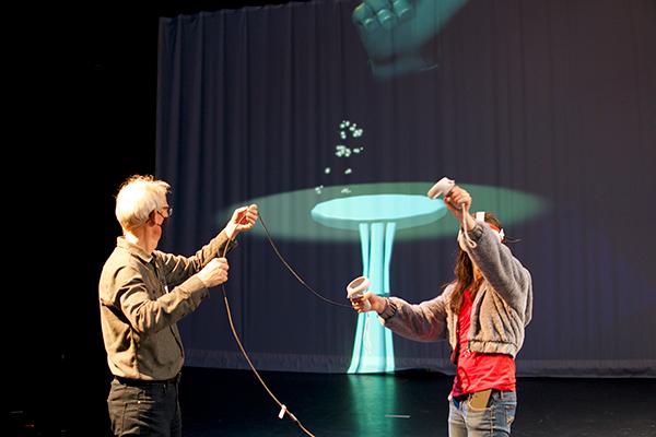A man holds the cord of a VR headset while a person in a red shirt and gray sweatshirt experiences a VR dance experience while the animation is projected onto a screen in the background