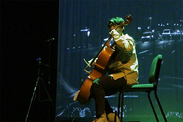 A man sitting on a chair playing a cello against a backdrop of a screen with a cityscape projected onto it.