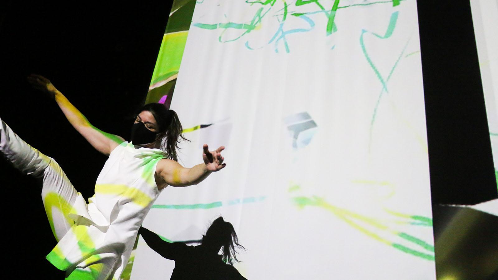 A dancer dressed white extends her arms in front of a white and green projection