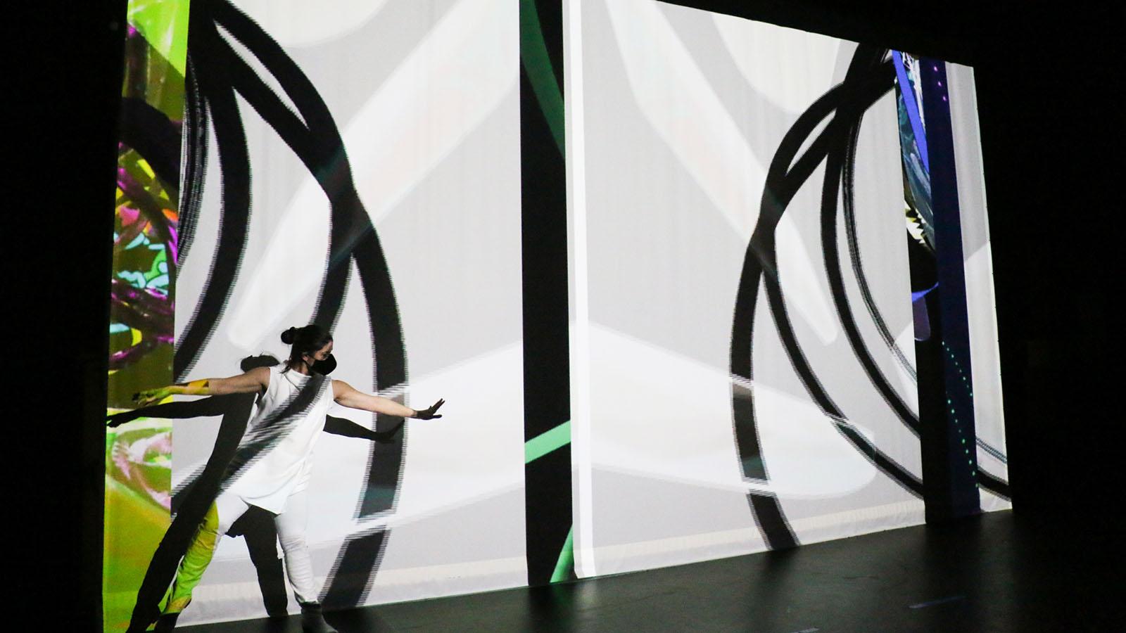 A swirling projection is layered onto of a dancer with spread legs and outstretched arms