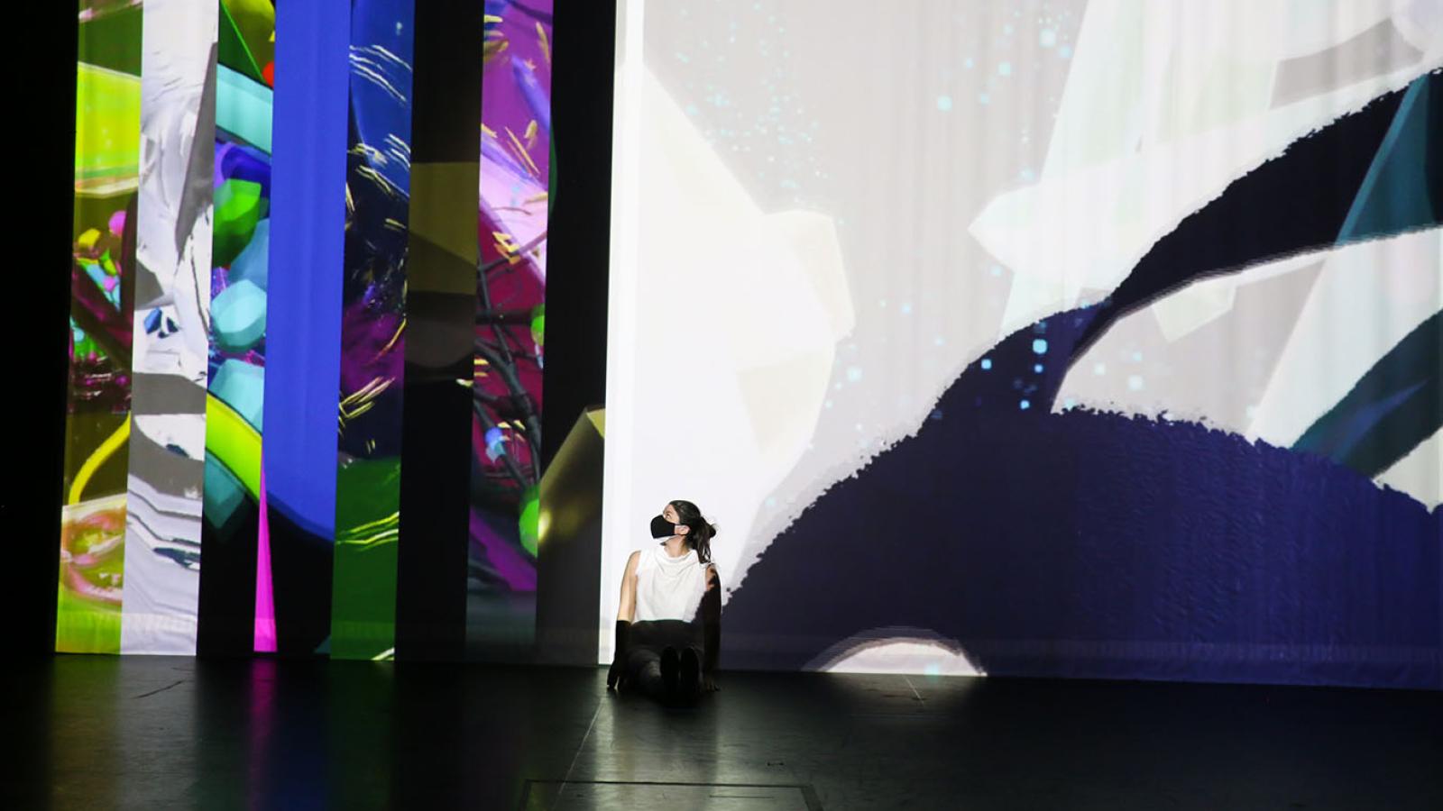 A dancer sits in front of a multicolored and layered image projection