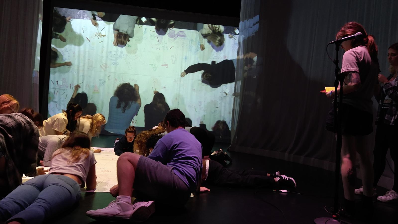 A group of students drawing on a large piece of white paper, while a live feed of their drawing is project on the wall behind them
