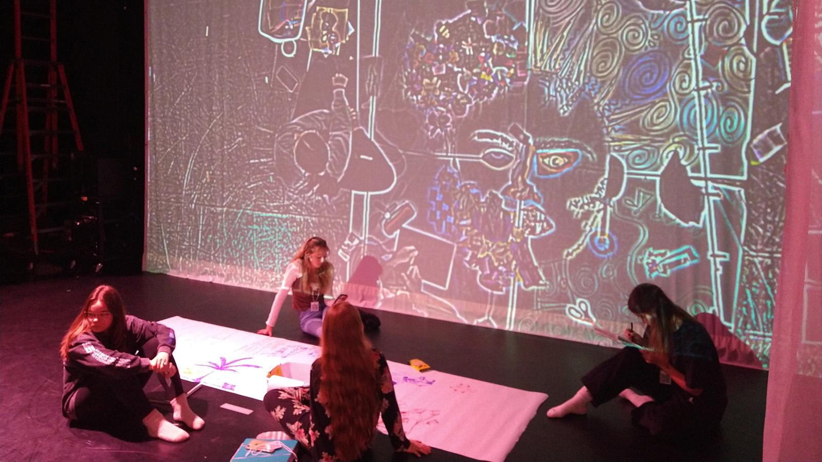 Four students drawing on a large piece of white paper, while a live feed of their drawing is project on the wall behind them