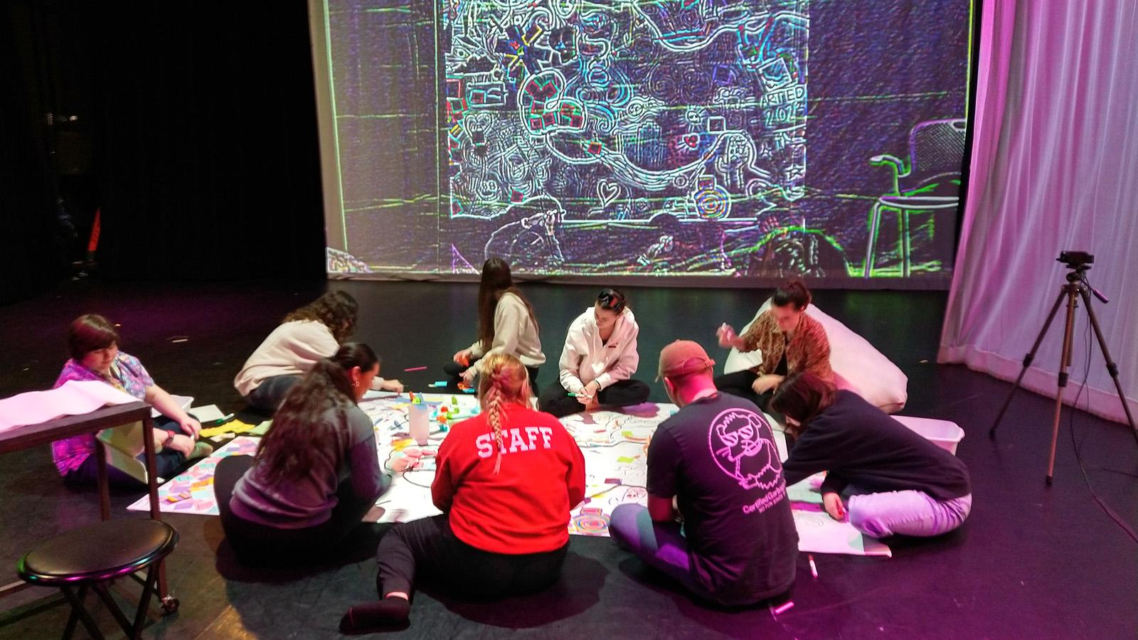 A large group of students sitting in the Motion Lab around a large piece of paper and drawing