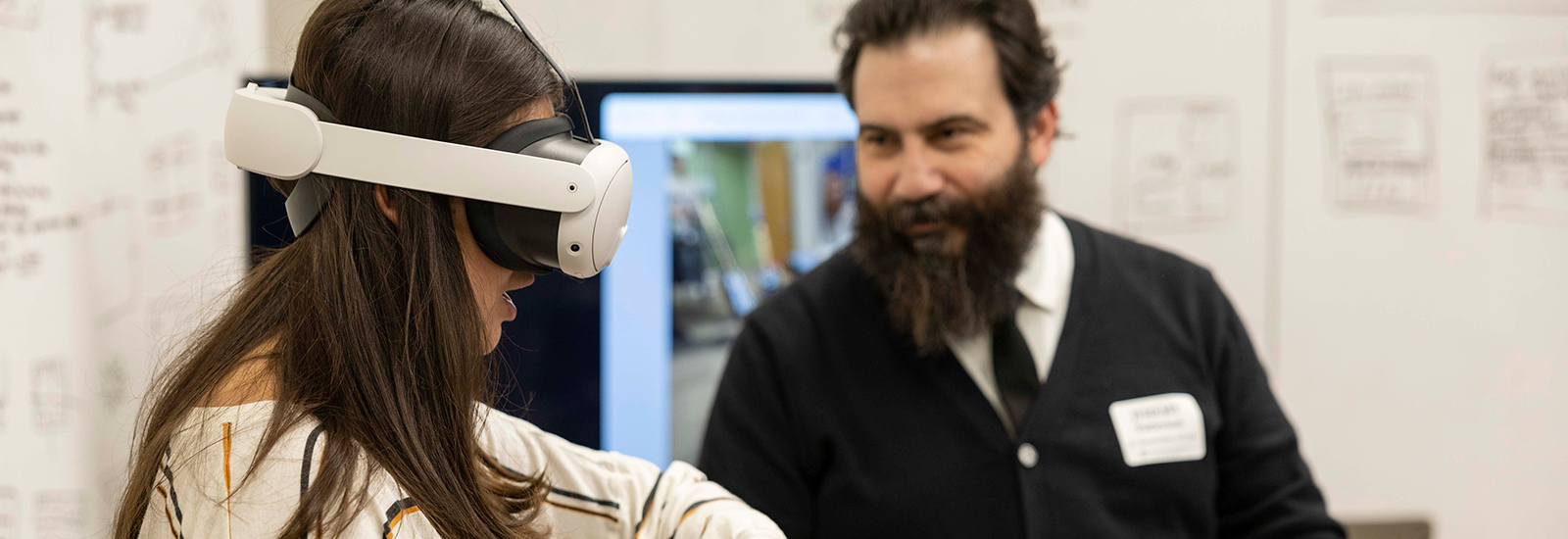 A woman stands in the foreground wearing a VR headset while a man helping to demo a project stands in the background, directing her.