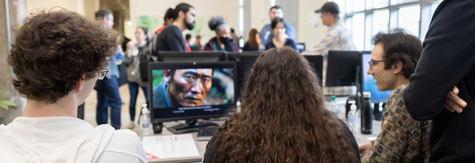 A group of three students sit at a table, watching a student-made video on a desktop computer. In the background, people socialize in the ACCAD computer lab during the Open House.